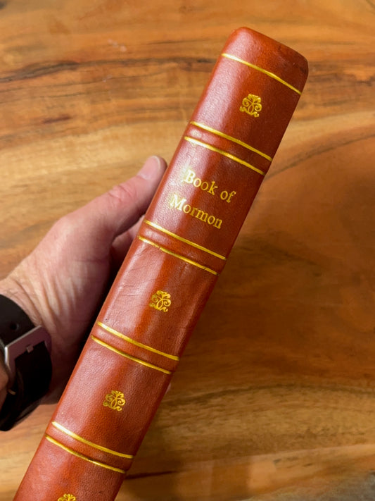 Replica 1869 leather-bound book titled 'Book of Mormon' held by a hand on a wooden surface