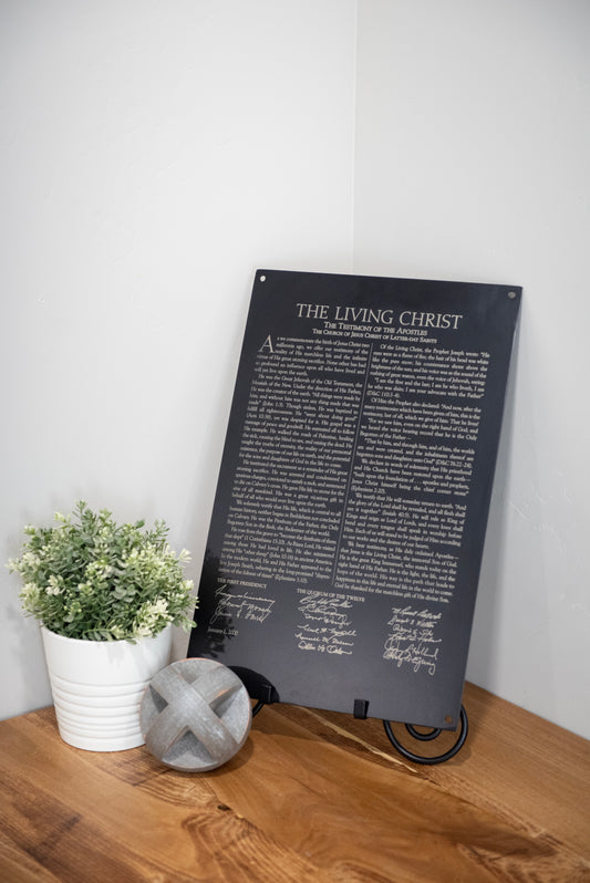 A black steel wall decor featuring text from 'The Living Christ' proclamation, displayed on a wooden surface with a white vase holding flowers to the left.