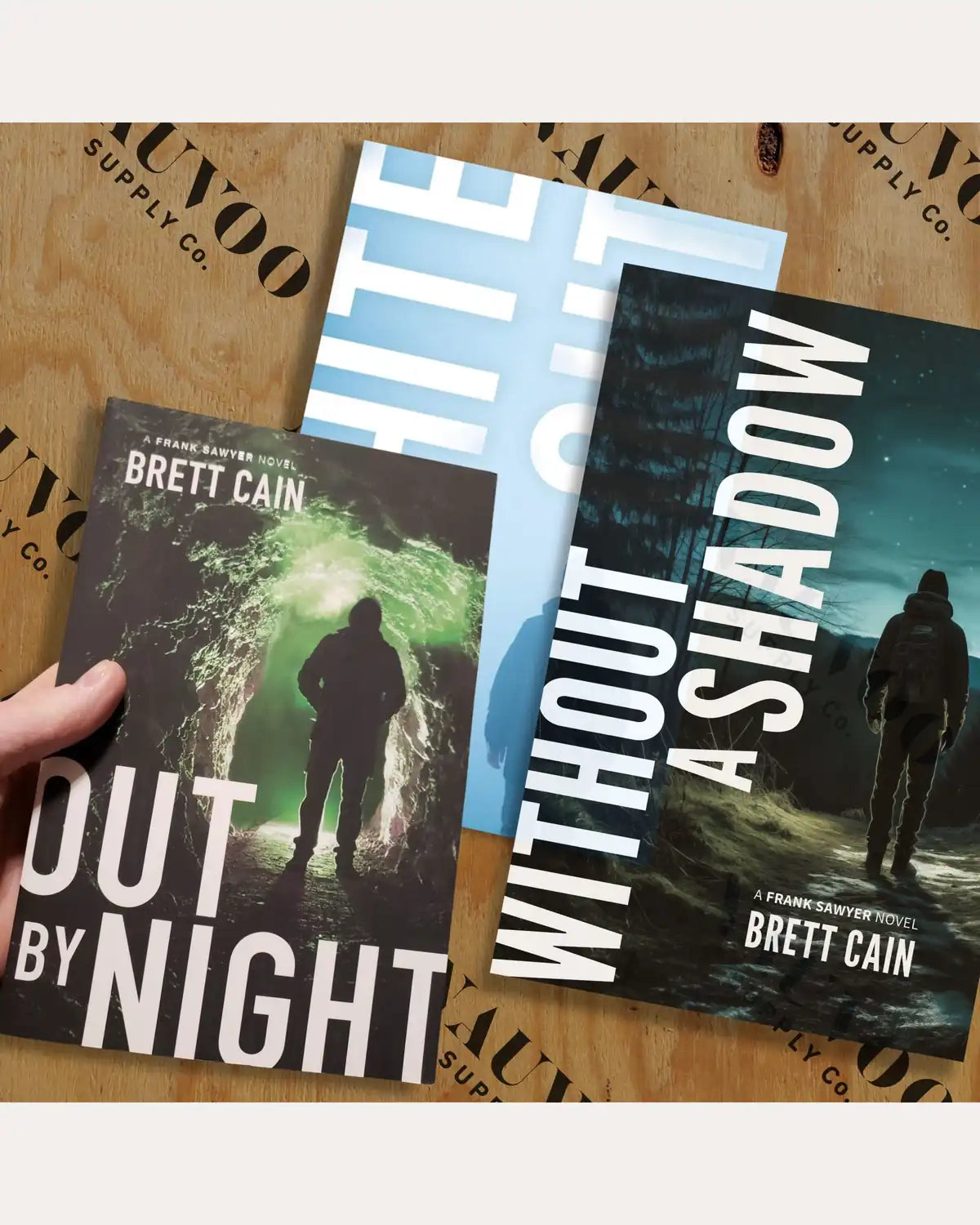 Three Frank Sawyer suspense novels by Brett Cain, Out by Night, Whiteout and Without a Shadow, fanned on a wooden crate.