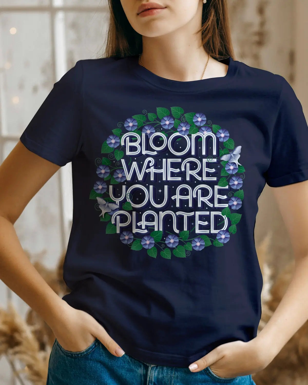 A navy t-shirt printed with "Bloom where you are planted," lettered inside a wreath of morning glories and luna moths.