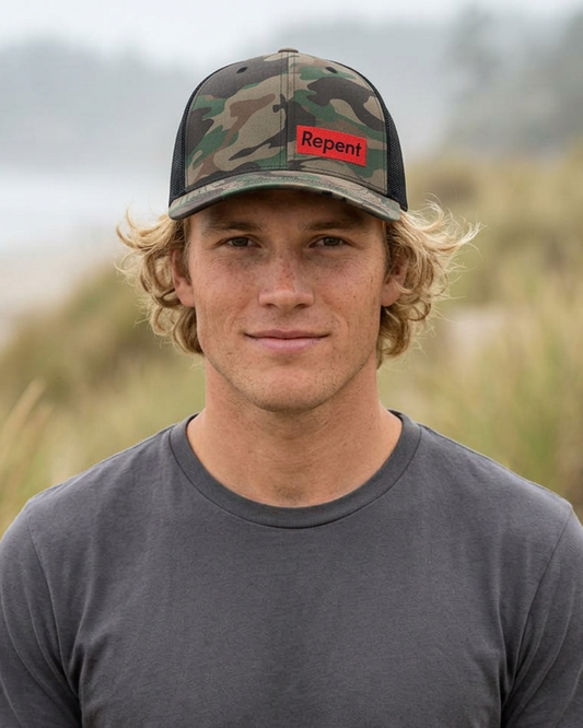 A Nauvoo Supply Co. product photo of a man on a beach dune wearing a camouflage trucker hat with a red 'Repent' patch.